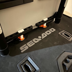 May include: A black and grey Sea-Doo storage container with multiple compartments and mounting brackets. The container has the Sea-Doo logo in silver and features orange and white accents. The interior is light grey.