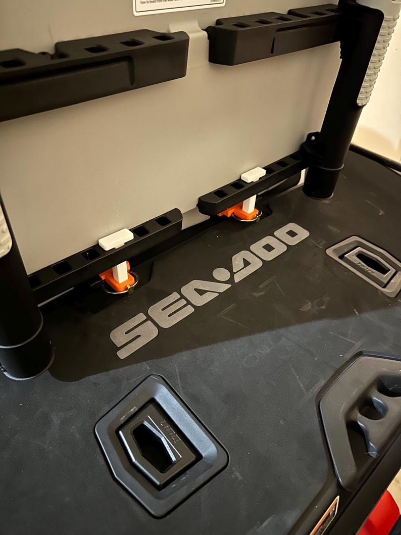 May include: A black and grey Sea-Doo storage container with multiple compartments and mounting brackets. The container has the Sea-Doo logo in silver and features orange and white accents. The interior is light grey.