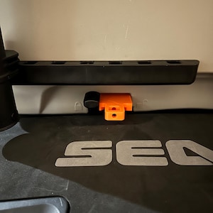 May include: A close-up of a black plastic component with an orange latch. The component has several rectangular openings and is mounted on a grey surface. The word "SEA" is visible in grey letters.