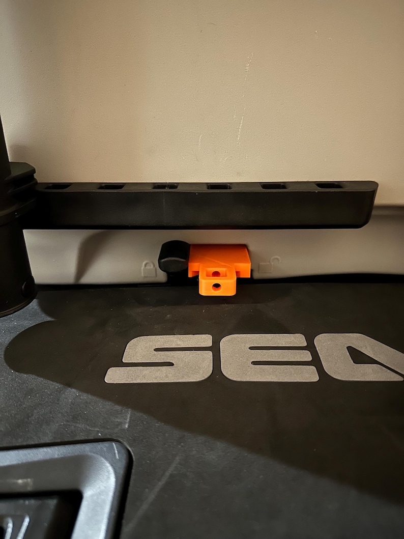 May include: A close-up of a black plastic component with an orange latch. The component has several rectangular openings and is mounted on a grey surface. The word "SEA" is visible in grey letters.