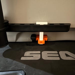 May include: A black plastic component with a series of holes, a white rectangular piece, and an orange and white bracket. The component is mounted on a surface with the letters "SEA" visible.