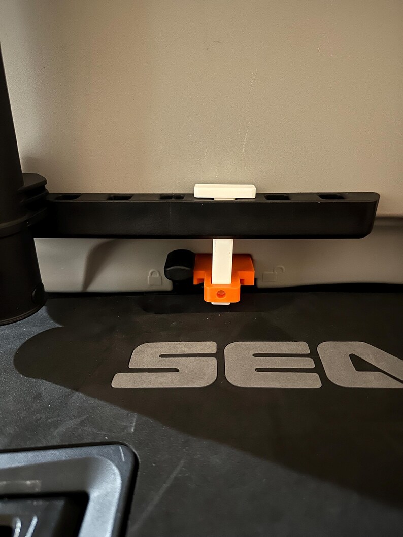 May include: A black plastic component with a series of holes, a white rectangular piece, and an orange and white bracket. The component is mounted on a surface with the letters "SEA" visible.