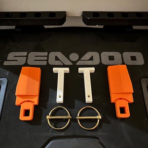 May include: A selection of Sea-Doo components, featuring two orange latches, two white T-shaped elements, and two gold-coloured split pins. The parts are displayed on a black surface with the Sea-Doo logo.
