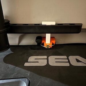 May include: A black plastic rack with multiple slots, a white support, and an orange and black component with a metal ring. The word "SEA" is visible in grey on a black surface.