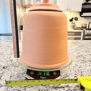 May include: A terracotta cloche with a lid, sitting on a glass plate atop a digital scale. The cloche is approximately 10 inches tall and 8 inches in diameter. A measuring tape and ruler are also in the frame.