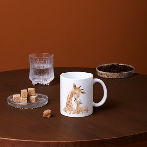 May include: White ceramic mug with a brown and white illustration of a mother giraffe and her baby giraffe. The giraffe's are surrounded by white flowers.