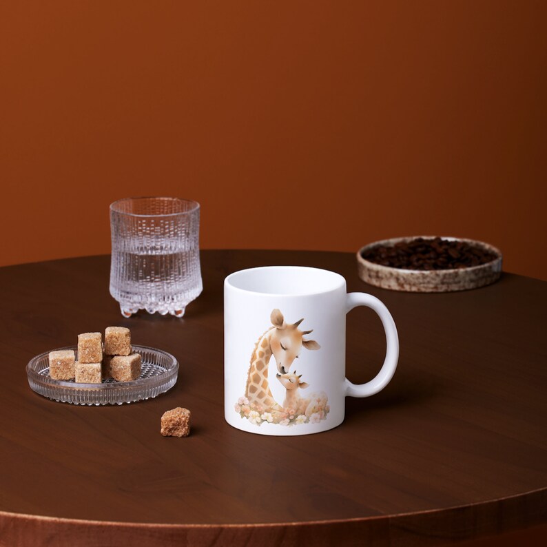 May include: White ceramic mug with a brown and white illustration of a mother giraffe and her baby giraffe. The giraffe's are surrounded by white flowers.