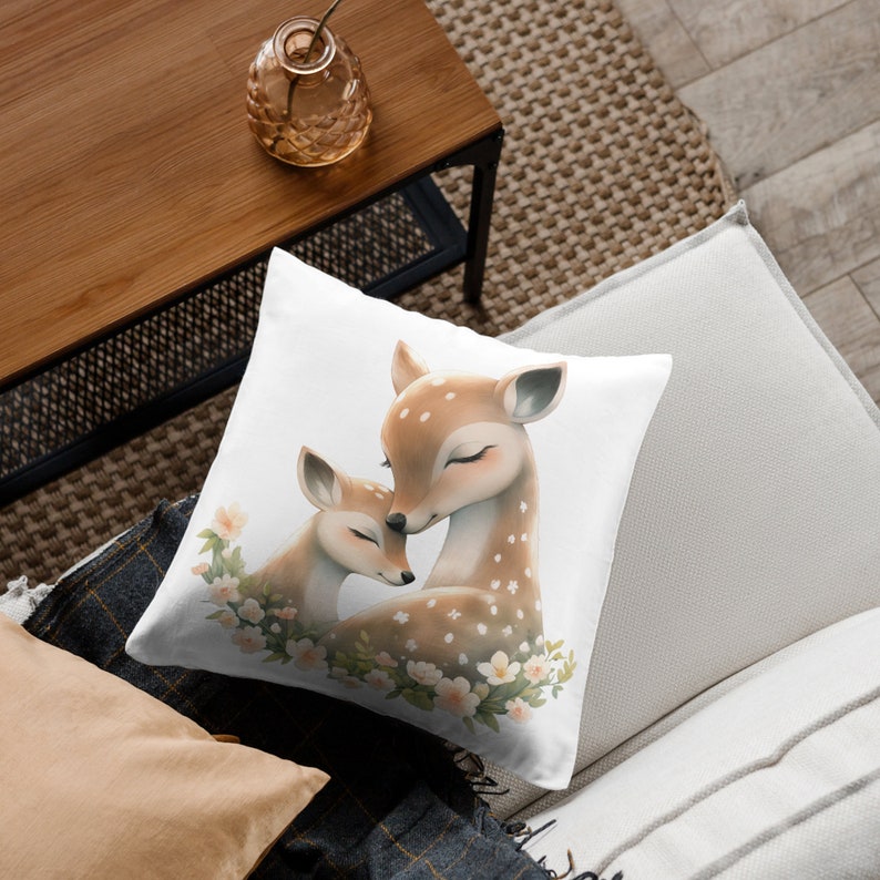May include: A white throw pillow with a watercolor illustration of a mother and baby deer. The deer are brown with white spots and are surrounded by white flowers.