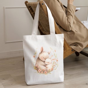 May include: A white canvas tote bag with a design of two brown rabbits hugging each other surrounded by pink flowers. The rabbits are in a circle of green leaves.