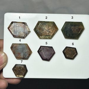 May include: A white display card featuring seven faceted, hexagonal-shaped gemstones labeled with numbers one through seven. The stones exhibit various earthy tones, including shades of brown, olive green, gray, and deep reddish-purple, with visible internal inclusions and natural color variations across each individual piece.