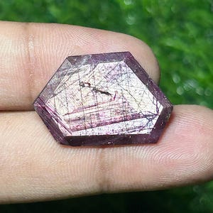 以下が含まれることがあります： 深いマゼンタと紫の色合いを持つ、六角形のファセットカットされた宝石。石の表面には、細かい暗いインクルージョンと線状の亀裂が複雑なネットワーク状に広がっており、質感のある多次元的な外観を作り出しています。