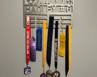 May include: A grey metal soccer medal display featuring the name "HADLEY" and the number "61". Below, ribbons and medals are suspended, including those for "INDY PIKE FEST FINALIST" and "SUPERSTAR".