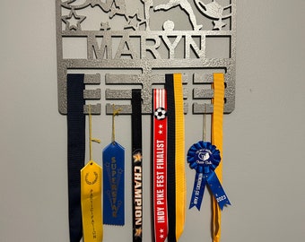 May include: A silver metal sports-themed medal display with the name "MARYN" cut out. The display features cut-out figures of athletes playing basketball, soccer, and other sports. Several medals and ribbons hang from the display, including ribbons that say "CHAMPION" and "SUPERSTAR".