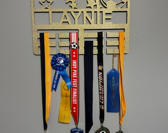 May include: A gold-coloured medal display featuring cut-out sports figures, stars, and the name "AYNIE." It holds several medals and ribbons, including ones that say "Cheer Club," "INDY PIKE FEST FINALIST," and "CHAMPION."