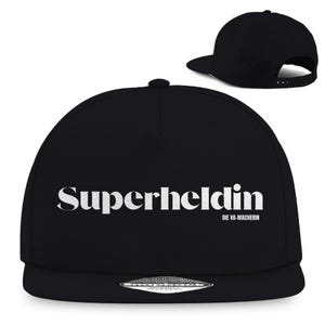 Könnte beinhalten: Schwarze Snapback-Kappe mit dem Wort "Superheldin" in weißen Buchstaben auf der Vorderseite. Darunter steht "DIE VA-MACHERIN". Die Kappe hat einen flachen Schirm und einen verstellbaren Druckknopfverschluss.