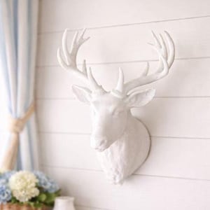May include: A matte white wall-mounted deer head sculpture featuring detailed antlers and a realistic facial structure. The piece is crafted with a smooth finish, capturing the form of a stag with ears pointed outward and a neck that tapers downwards.