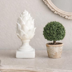 May include: A white artichoke ornament and a small potted topiary sit on a light-coloured, ornate table. A round, cream-coloured mirror with a decorative frame hangs on the wall behind the table. The scene has a soft, neutral colour palette.