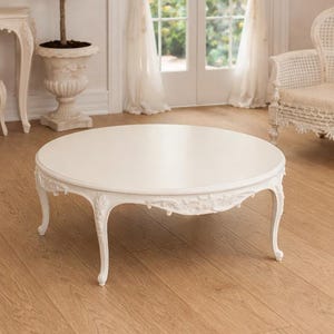 Op de afbeelding: Een ronde, witte salontafel met gebogen poten en gedetailleerd houtsnijwerk langs de rand. De tafel heeft een glad, cirkelvormig tafelblad en is afgewerkt met een effen witte lak, rustend op vier slanke, sierlijke poten.