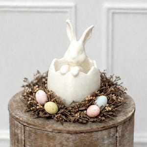 Könnte beinhalten: Eine weiße Keramik-Osterhasenfigur, die aus einem zerbrochenen Ei kommt, umgeben von einem Kranz aus getrocknetem Laub und pastellfarbenen Eiern. Das dekorative Stück steht auf einem kleinen, verwitterten Beistelltisch aus Holz mit einer Schublade.