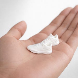 May include: A pair of miniature white sneakers, featuring a classic athletic design. The shoes are detailed with laces and a subtle logo, presented on a neutral background. The sneakers are small, likely for display or as a collectible item.