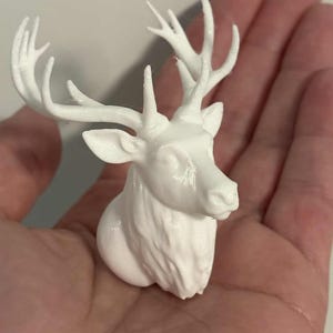 Puede incluir: Una pequeña figura de cabeza de ciervo impresa en tres dimensiones de color blanco, con astas detalladas, orejas puntiagudas y un cuello texturizado. El objeto está esculpido con un acabado liso y representa a un ciervo mirando al frente.