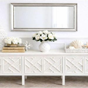 May include: A long white wooden console table with six cabinet doors featuring a geometric lattice pattern. On top sits a white patterned ceramic jar, a stack of two books, a white floral arrangement in a vase, and a tray with seashells.