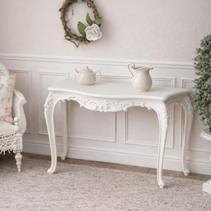 May include: A white, ornate table with curved legs and a decorative edge. A teapot and a creamer sit on the table. A floral wreath hangs on the wall above the table. The room has a vintage, elegant aesthetic.