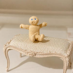 May include: A small, tan clay figure with blue eyes sits on a miniature, ornate bench. The figure has its arms outstretched. The bench has a cream-coloured, patterned seat and a carved, off-white frame.