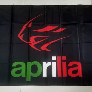 May include: Black flag featuring the Aprilia logo. The logo includes a red stylized design above the word "aprilia" in green, white, and red. The flag has grommets for hanging.