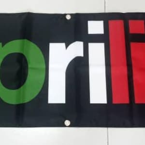 May include: A black banner with the word "aprilia" in green, white, and red letters. The letters are in a stylized font and the word is centered on the banner. There is a registered trademark symbol in the upper right corner of the banner.