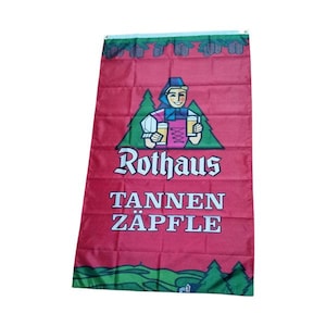 Op de afbeelding: Een rode vlag met een groene rand en een wit logo. Het logo toont een cartoonfiguur dat twee bekers bier vasthoudt. De tekst "Rothaus Tannen Zapfle" is onder het logo gedrukt.