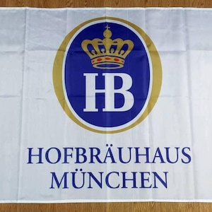 Könnte beinhalten: Weiße Flagge mit einem blauen und goldenen Logo. Das Logo zeigt eine Krone und die Buchstaben "HB" in einem Kreis. Der Text "HOFBRAUHAUS MÜNCHEN" ist unter dem Logo gedruckt.