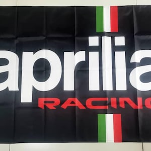 Può includere: Bandiera nera con testo bianco che recita "aprilia racing" e un design di bandiera italiana rosso, bianco e verde.