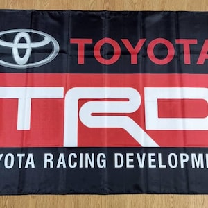 Op de afbeelding: Zwarte vlag met een rood en wit Toyota TRD-logo. Het logo zegt "Toyota Racing Development" in witte tekst.