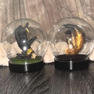 May include: Two clear glass globes, each housing a hand sculpture. One is black with gold detailing, the other gold. They sit on black bases, possibly for display. A unique decorative or artistic item.