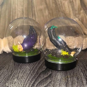 May include: Two glass globes, each with a claw sculpture. One is purple, the other black with green details. Both sit on green moss and stones, atop a black base. The globes are decorative items.