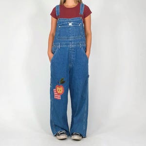 May include: Blue denim overalls with an apple patch on the left leg. The overalls have adjustable straps, pockets, and a wide-leg design. The apple patch features a red apple with green leaves and a red and white checkered square.