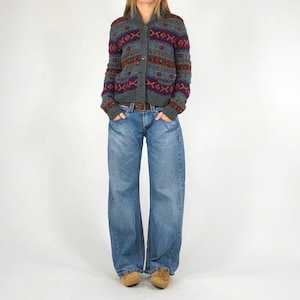 May include: A gray cardigan with a geometric pattern in shades of red, teal, and brown. The cardigan has a shawl collar and button closure. The person is wearing light blue, wide-leg jeans and brown shoes.