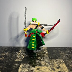 May include: A handcrafted pipe cleaner figurine depicting a character with green hair, wearing a green coat with yellow accents and black trousers. The figure holds two swords in its hands and has a third sword held horizontally in its mouth.