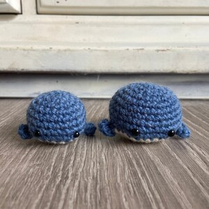 May include: Two crocheted whale plushies, one larger than the other, both in a blue color with black eyes. The whales are sitting on a wooden surface.
