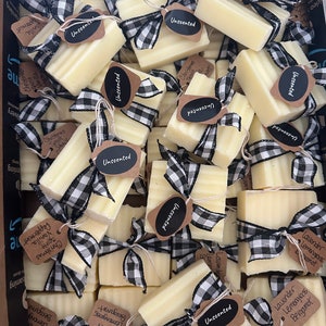 May include: A close-up of a variety of handmade soap bars wrapped in black and white checkered ribbon with brown paper tags. The tags are labeled with different scents, including "Unscented", "Lavender Lemongrass Bergamot", and "Christmas Spirit Vanilla Peppermint".