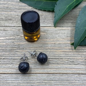 May include: A pair of black lava stone earrings with silver posts. The earrings are on a wooden surface next to a small brown glass bottle with a black lid.