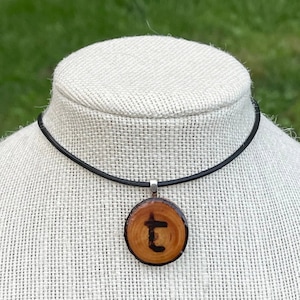 May include: A round wooden pendant necklace with a black leather cord. The pendant features a natural wood grain design with the letter "t" in black. The necklace is displayed on a white mannequin head.