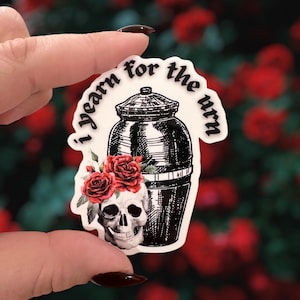 May include: A sticker featuring a black urn design with a skull adorned with red roses. The text "I yearn for the urn" is arched above the urn. The sticker is held up against a blurred background of red flowers.