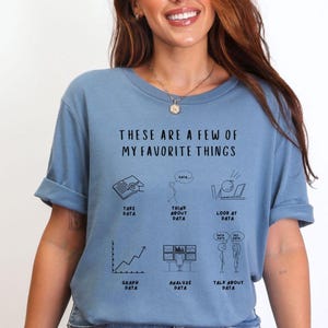 Può includere: T-shirt azzurra con la scritta "THESE ARE A FEW OF MY FAVORITE THINGS" e illustrazioni relative ai dati. La maglietta presenta disegni di un libro, una nuvoletta, un laptop, un grafico, monitor di computer e due persone che parlano.
