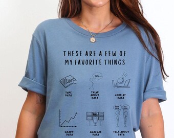 May include: A pale blue t-shirt with the text "THESE ARE A FEW OF MY FAVORITE THINGS" and data-themed graphics. The design includes images of a book, thought bubble, laptop, graph, computer screens, and two people conversing about data.