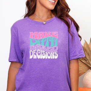 Könnte beinhalten: Ein lila T-Shirt mit dem Text "MAKE DATA BASED DECISIONS" in einem Retro-Schriftzug. Das Shirt ist ein kurzärmeliges Rundhals-Modell. Das Model trägt weiße Shorts. Das Shirt ist aus einem weichen, bequemen Material.