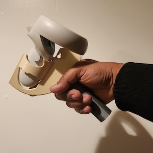May include: A beige 3D printed wall mount for a VR headset controller. The controller is white and has a black strap. The mount is designed to hold the controller in a vertical position.
