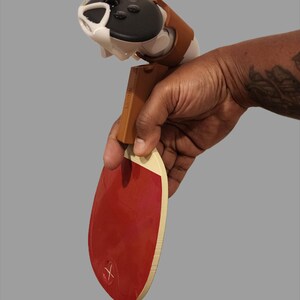 May include: A red ping pong paddle with a wooden handle and a white edge. The paddle is held in a hand, with a black and white device attached to the handle. The device has a brown strap.