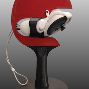 May include: A white video game controller is mounted on a black stand with a red, semi-circular base. The stand has a USB port and two buttons on the base.